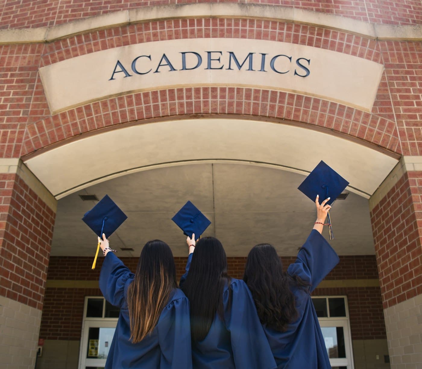 Three graduates raising their caps beneath an arch marked Academics